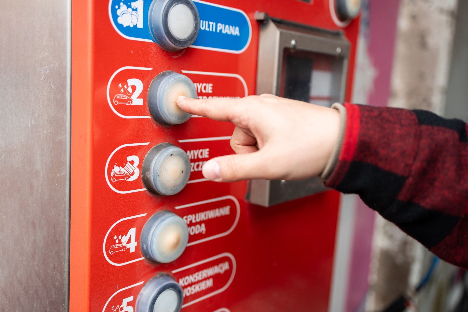 Self service car wash station featuring a red and white theme, a control panel, blue hose, instructional icons, and trees in the background under daylight.